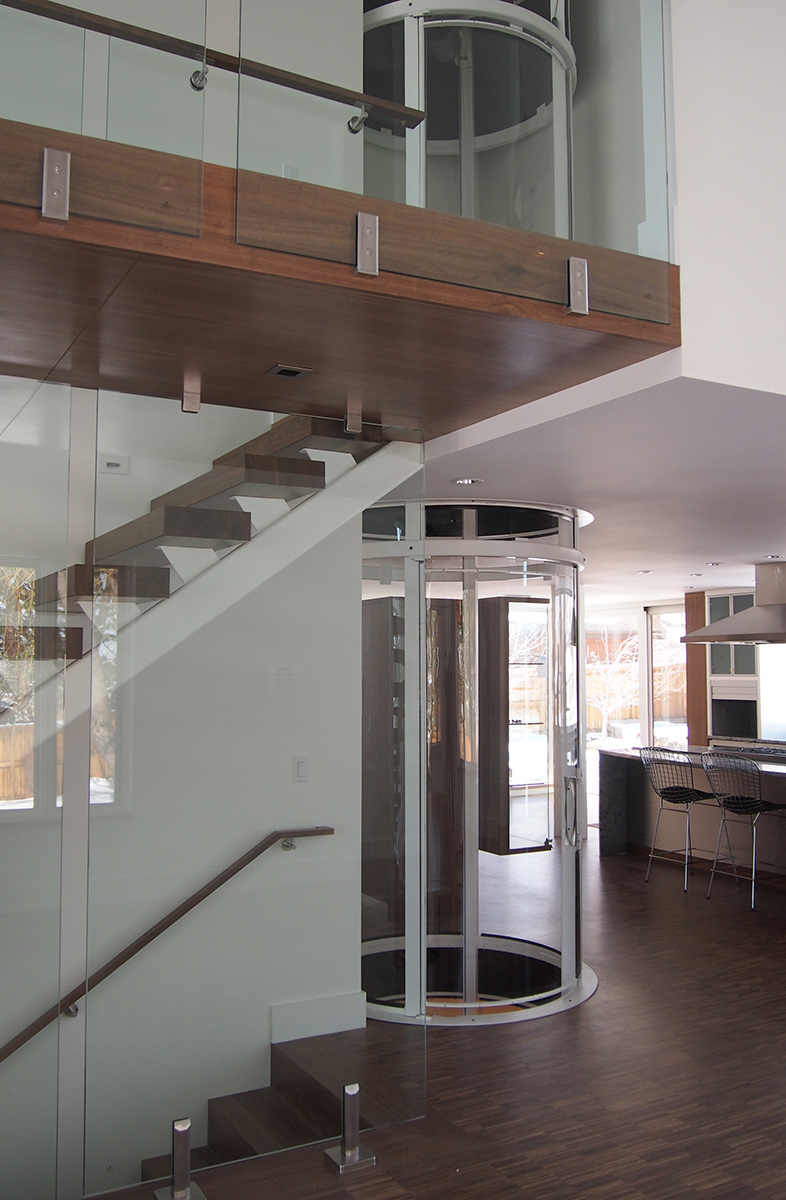 A round glass residential elevator spanning two floors beside an open staircase in a modern home