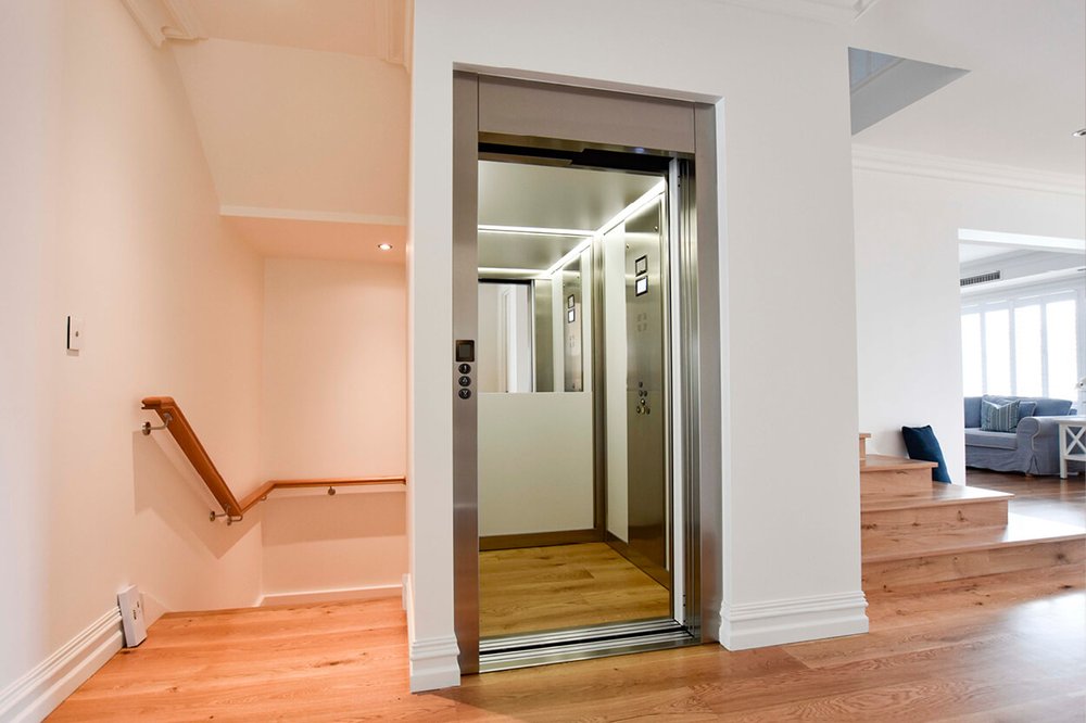 A home elevator with open doors and a lit cab beside the stairs in a bright private house